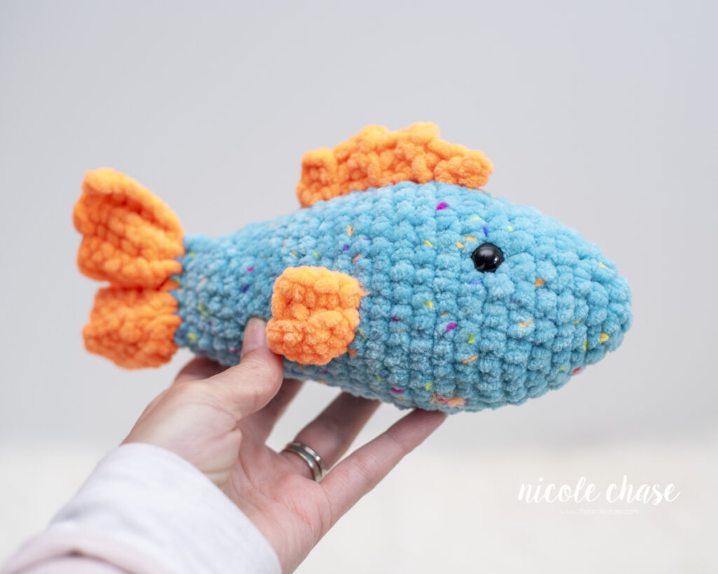 A hand holds a plush, larger version of the Finley the Fish amigurumi toy against a light background to show its size. This version is crocheted using a thick, textured blue blanket yarn with colorful flecks, featuring chunky orange plush fins and a matching tail.
