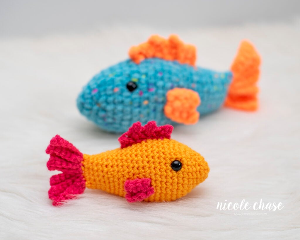 Two crocheted fish made from the Finley the Fish crochet pattern resting on white fur. In the foreground, an orange crochet fish has bright pink fins and tail. In the background, a blue flecked yarn fish is styled with bright orange fins and tail.