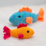 A vibrant orange and pink amigurumi fish sitting in the foreground on a white faux fur surface, with a blue and orange crocheted fish softly blurred in the background. This free fish crochet pattern features textured fins and safety eyes.