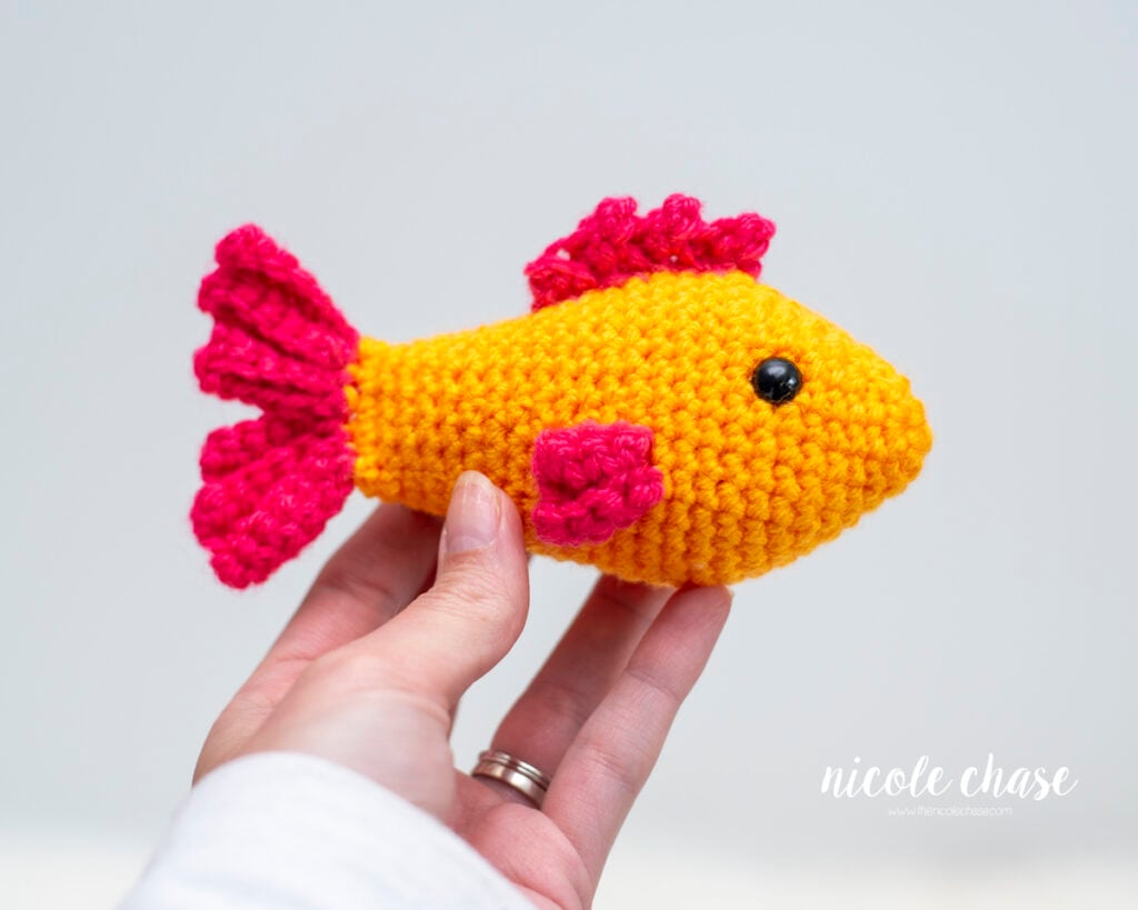 A hand holds up a finished Finley the Fish amigurumi toy to show its size against a plain, light background. The small, bright orange crocheted fish features textured pink fins on its side and top, a matching pink wavy tail, and black safety eyes.
