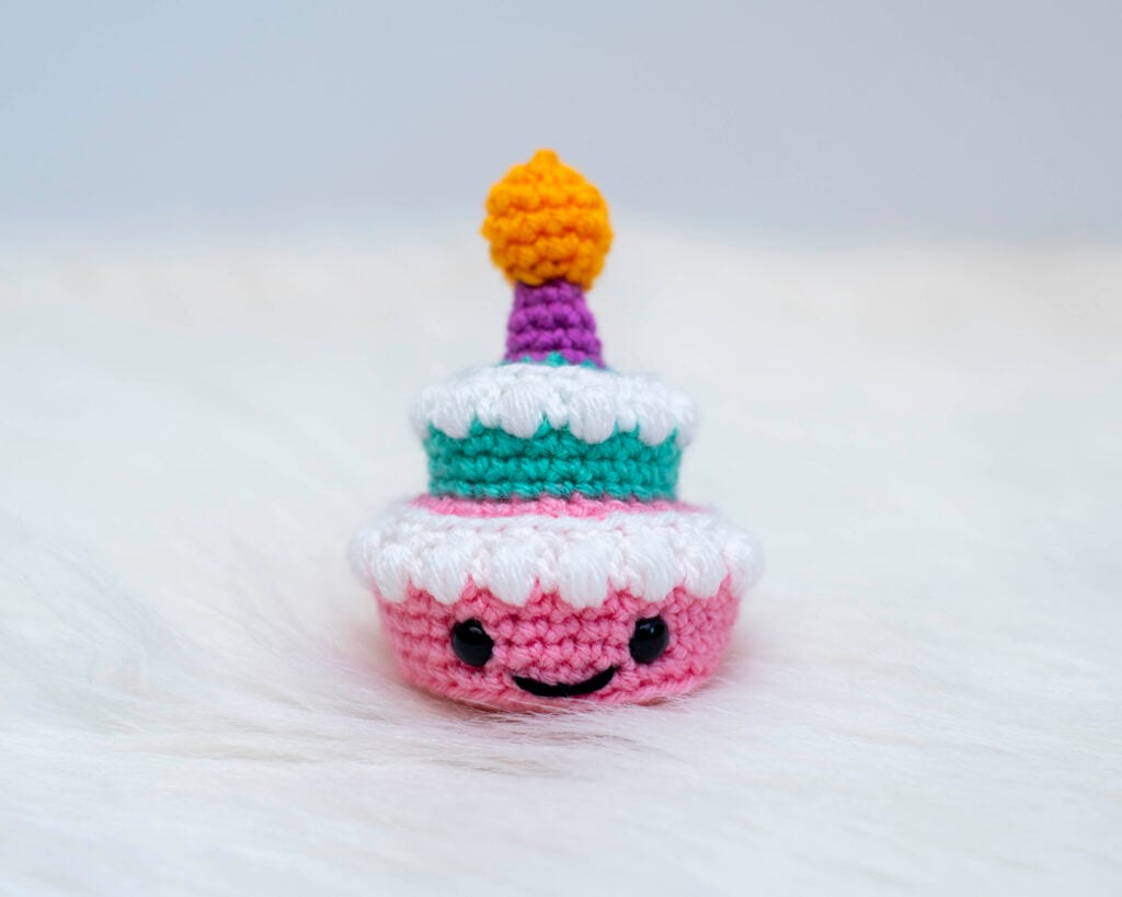 A close up of a small two-tier amigurumi birthday cake. The bottom tier is pink with an embroidered smile and safety eyes, topped with textured white frosting. The top tier is teal, also with textured white frosting. A purple candle with yellow flame is crocheted on top. The cake sits on a white, fluffy surface against a light background.