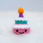 A close up of a small two-tier amigurumi birthday cake. The bottom tier is pink with an embroidered smile and safety eyes, topped with textured white frosting. The top tier is teal, also with textured white frosting. A purple candle with yellow flame is crocheted on top. The cake sits on a white, fluffy surface against a light background.
