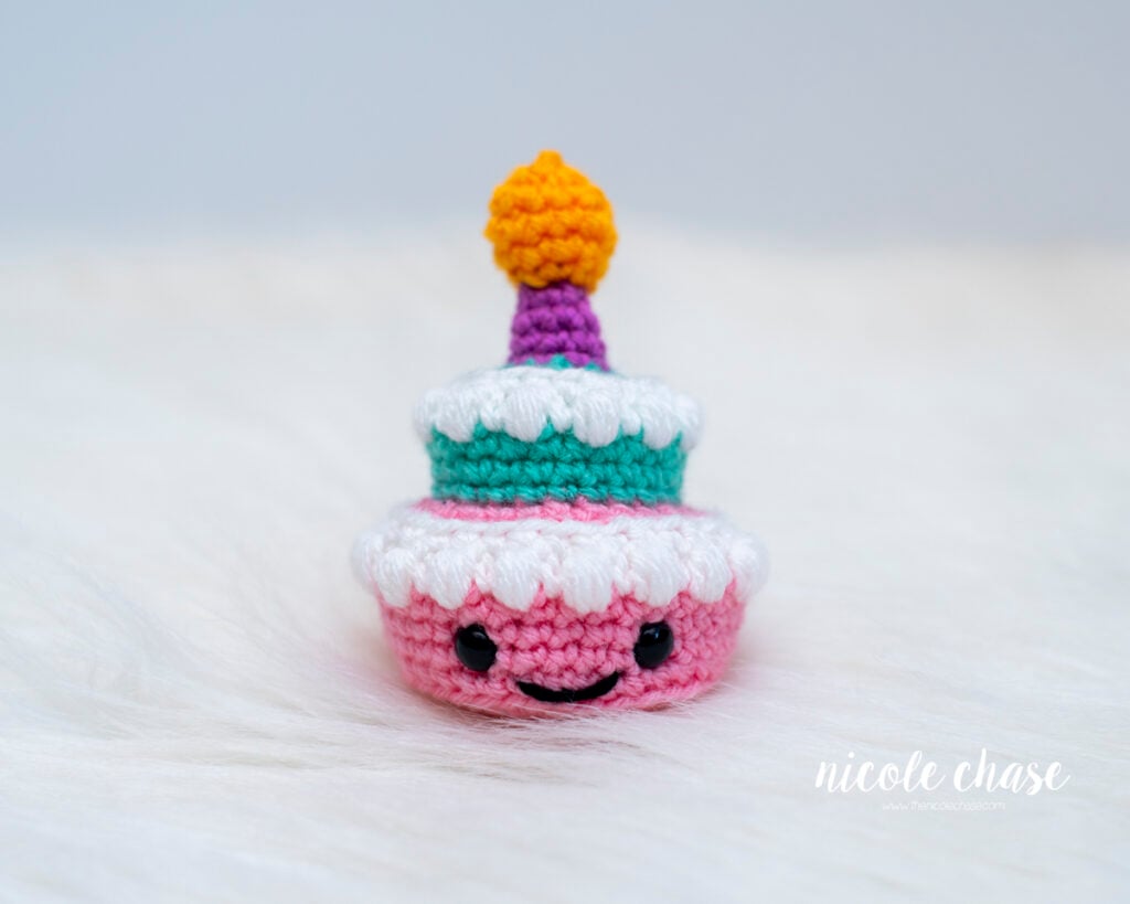 Small two-tiered amigurumi birthday cake crocheted in pink and teal yarn with white textured frosting, safety eyes, a smiling face, and a purple candle with a yellow flame.