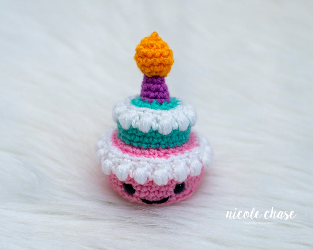 High-angle view of a small two-tiered amigurumi birthday cake made with pink, teal, white, purple, and orange yarn. The crocheted cake features white bobble stitch frosting on each tier, safety eyes, a stitched smile, and a candle topped with a flame, sitting on a soft white fur surface.