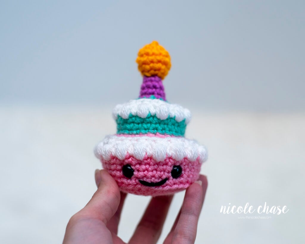 A hand holding a finished small two-tiered amigurumi birthday cake to show scale. The crocheted cake features a pink bottom tier with safety eyes and an embroidered smile, a teal top tier, white bobble frosting details, and a purple candle with an orange flame.