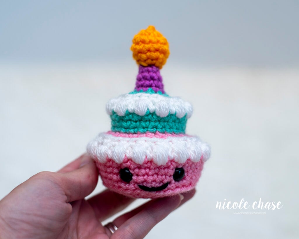 A hand holding a finished small two-tiered amigurumi birthday cake. The crocheted cake features a pink bottom tier with safety eyes and an embroidered smile, white puff stitch frosting, a teal top tier, and a purple candle topped with a yellow flame.