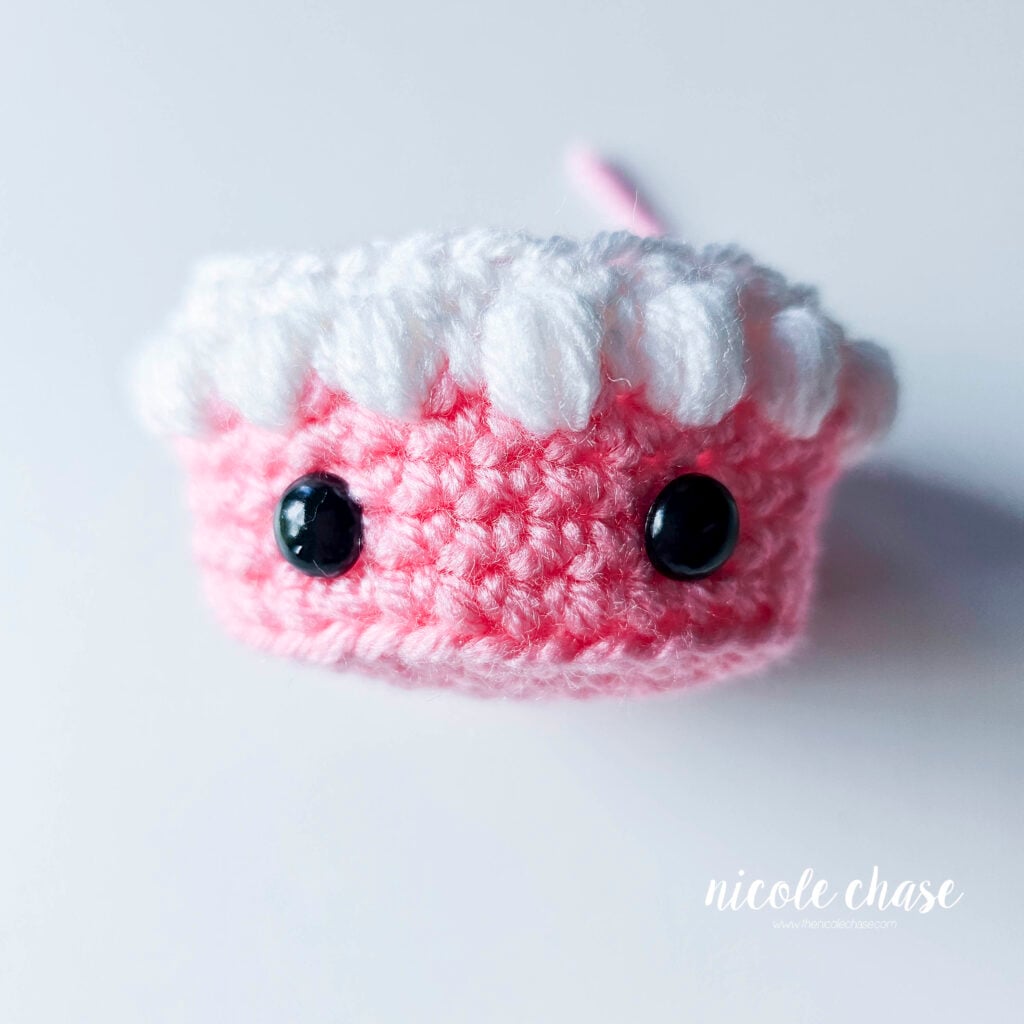 The black safety eyes have been inserted into the first tier of the crochet birthday cake, demonstrating the proper placement.