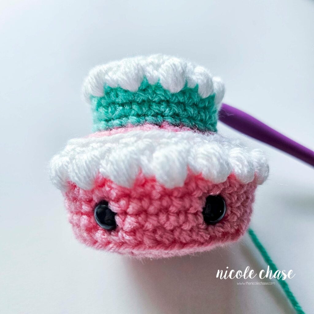 The second tier of the birthday cake crochet pattern has been completed in teal yarn with white puffy icing stitches on top of the pink base.