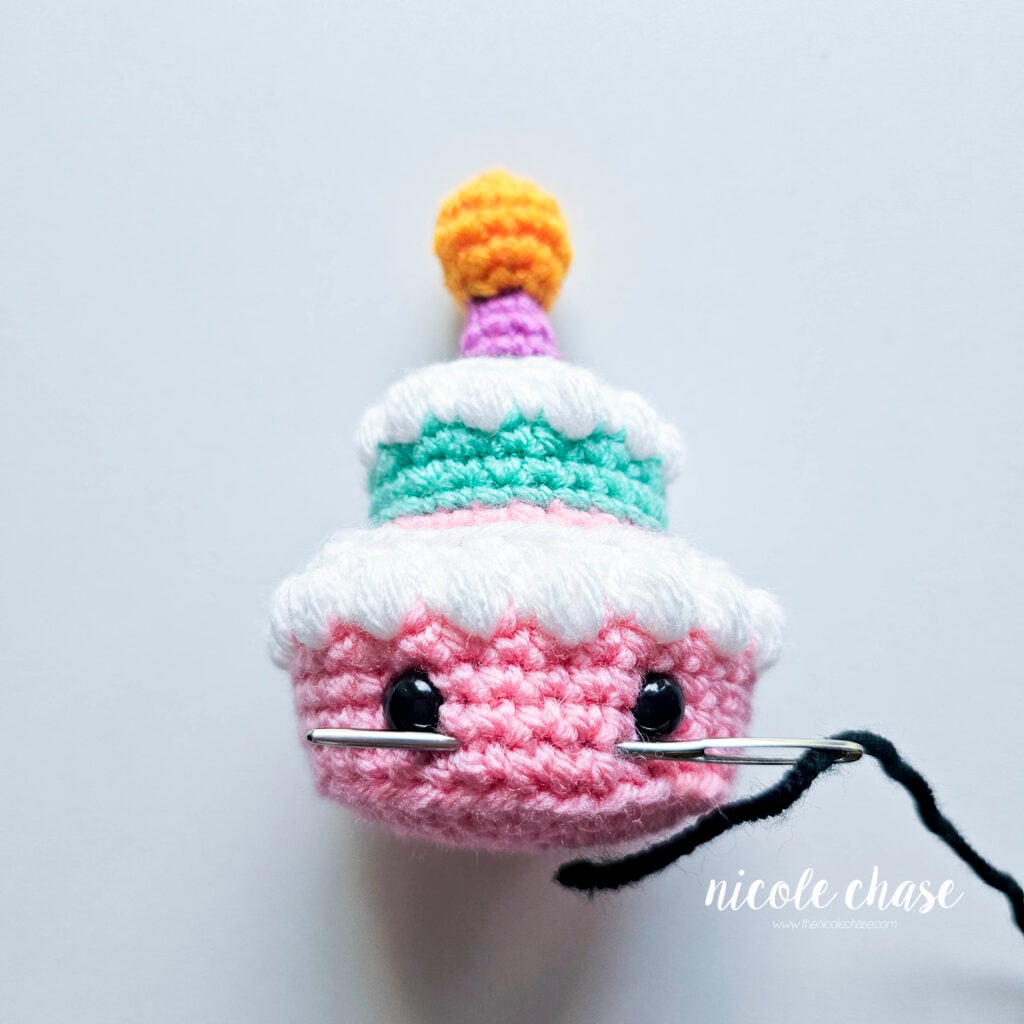 The first step in an embroidering a smile on the first tier of the crochet birthday cake is inserting your yarn needle through the desired stitches.