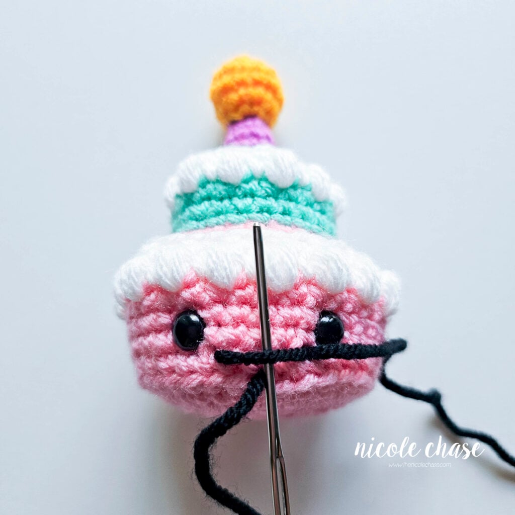 The yarn needle is pushed under the straight line across, preparing to loop over to create the dip in the smile.