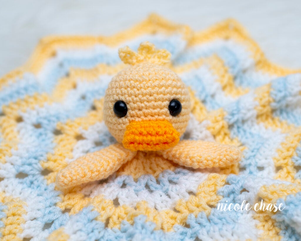 Close-up view of a yellow crocheted duck lovey showing the black safety eyes, small orange bill, and soft star blanket base.