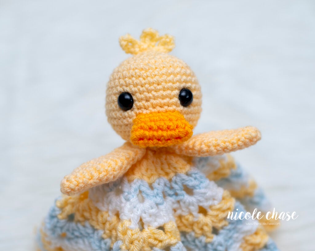 Front-facing close-up of the completed free duck lovey crochet pattern showing the head, outstretched wings, orange bill, and draped star blanket.