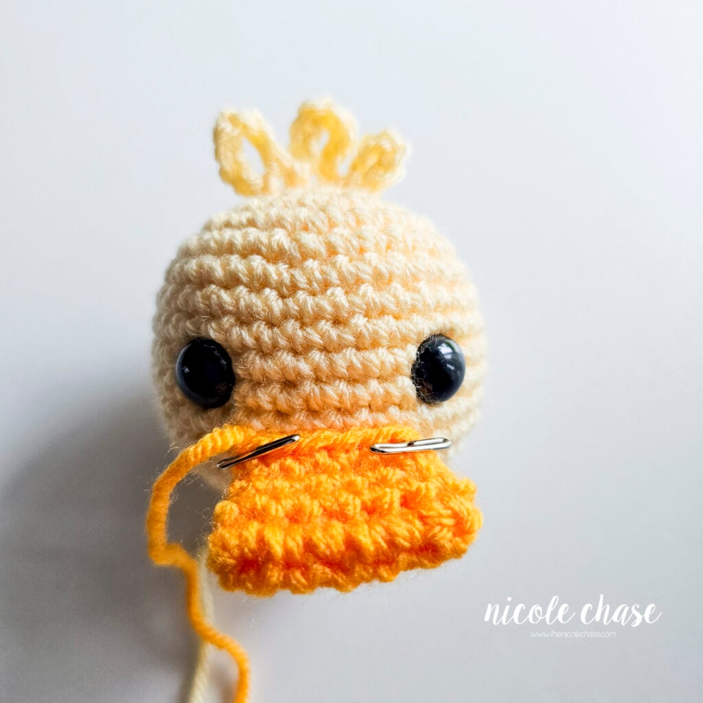 Orange crocheted duck bill pinned securely between the safety eyes on the yellow head before stitching.