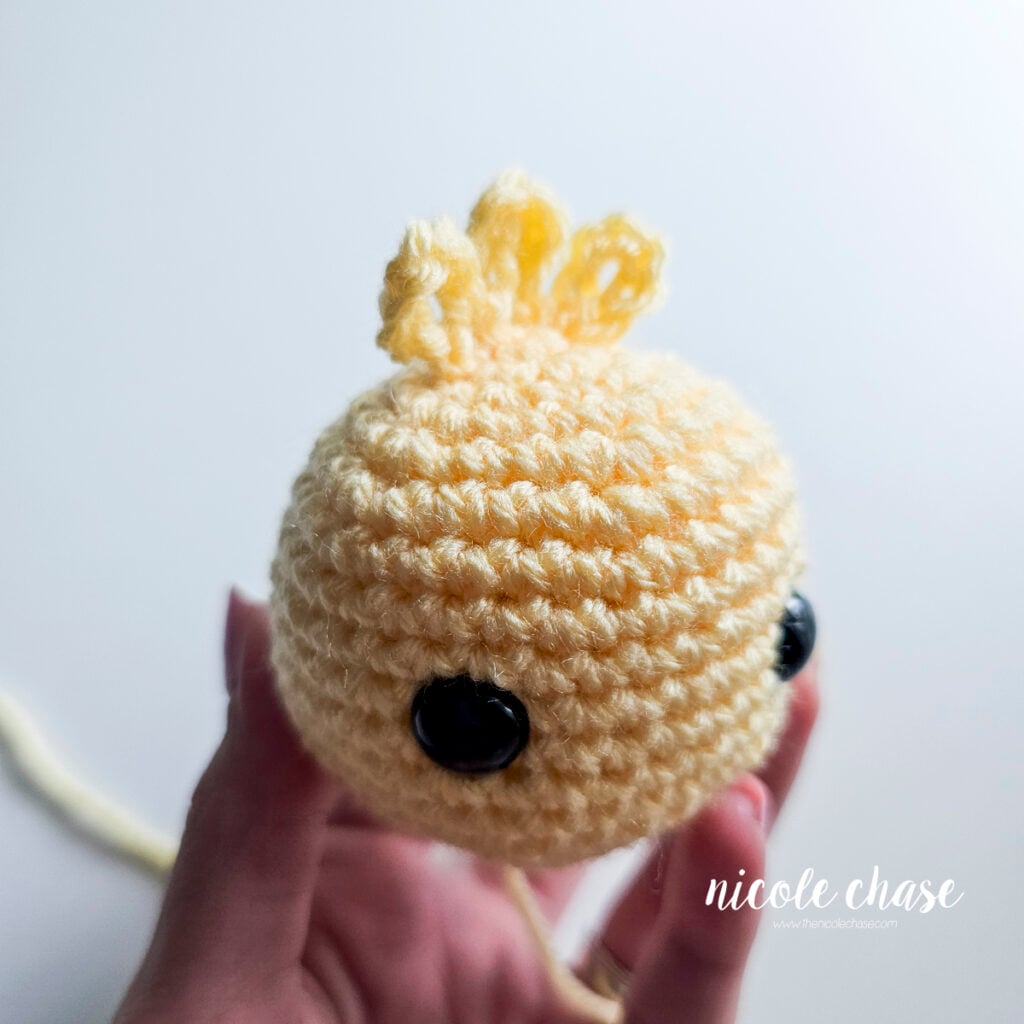 Side view of the yellow crocheted head with the feather tuft securely sewn centered between the safety eyes.