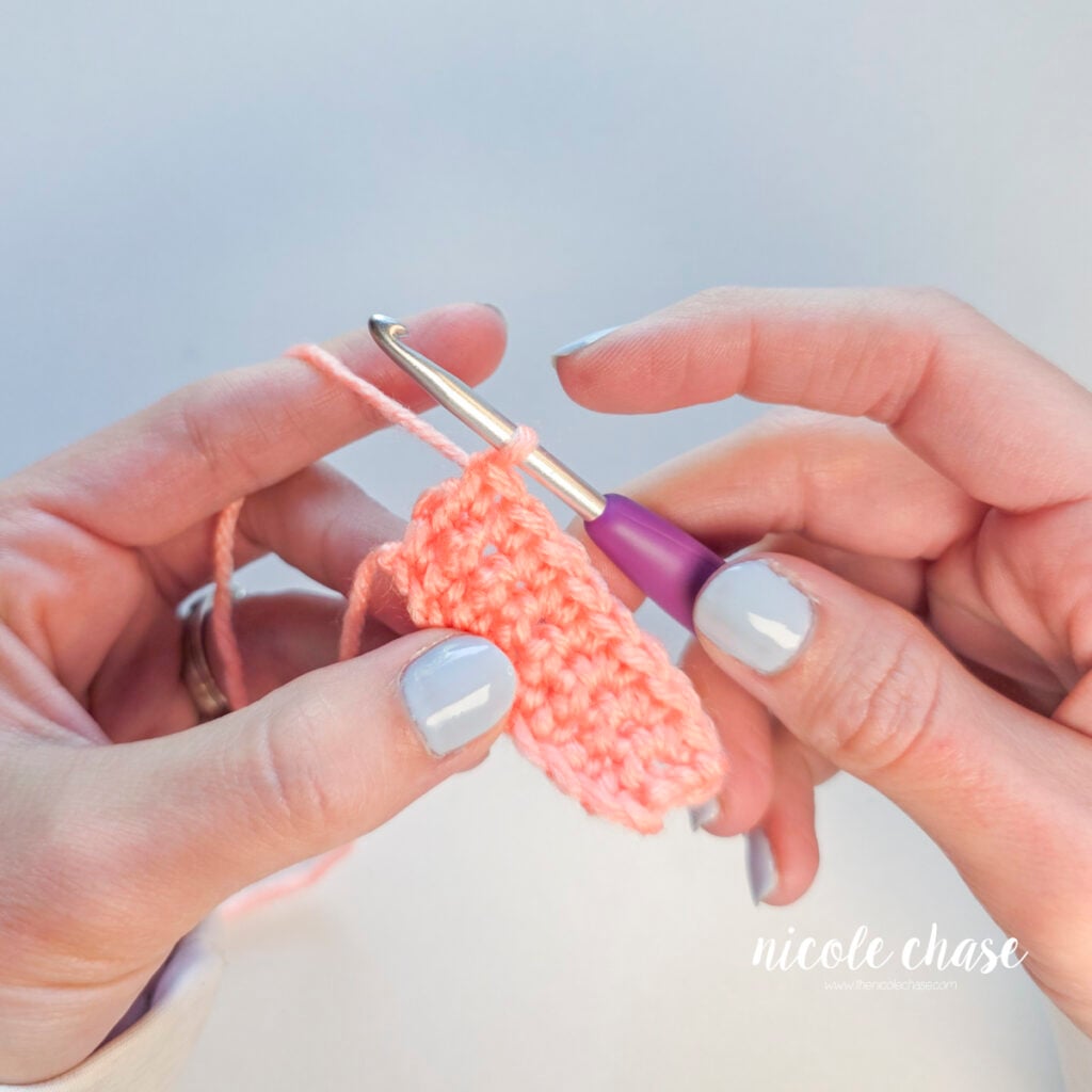 Hands holding a crochet hook with a single pink yarn loop on the hook at the end of a pink single crochet swatch.