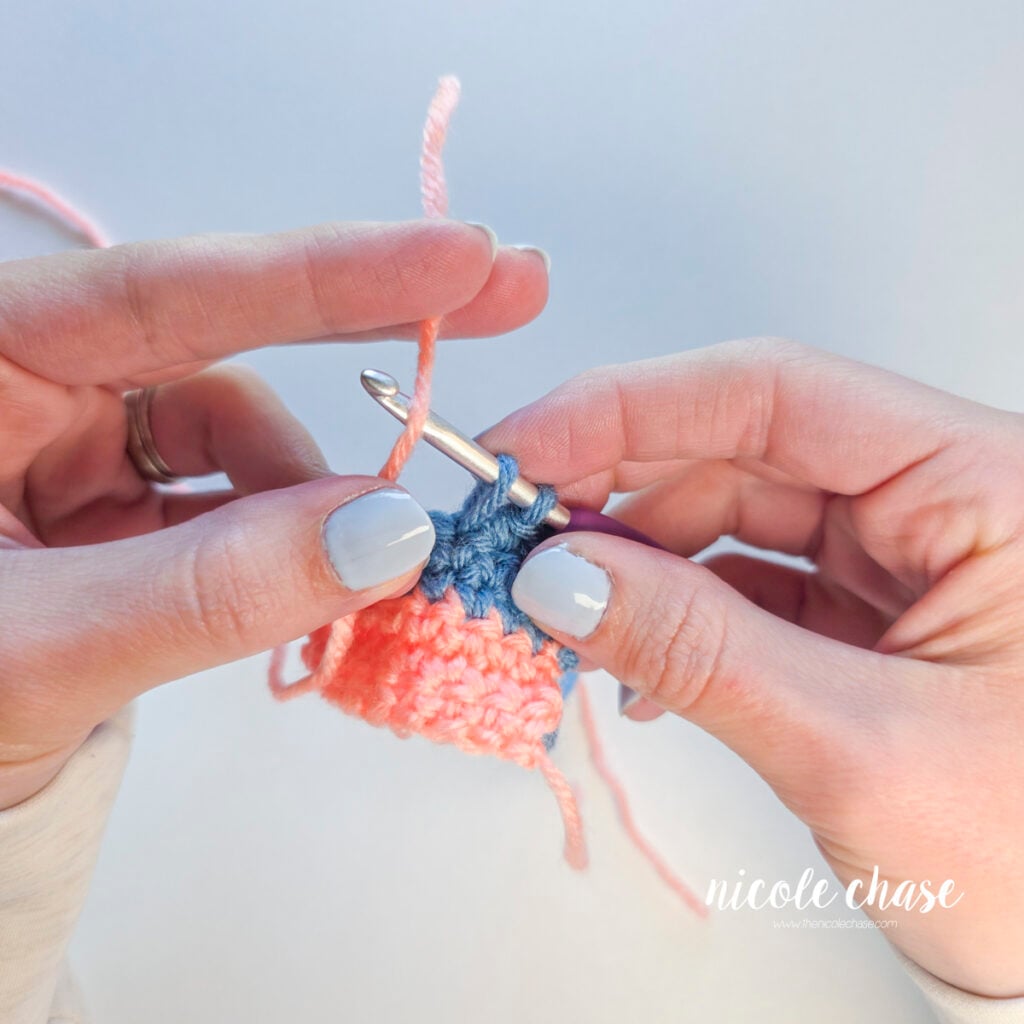Yarning over with pink yarn on a crochet hook containing two blue loops.