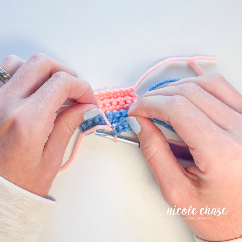 Hands tying pink and blue yarn tails together behind a crochet swatch.