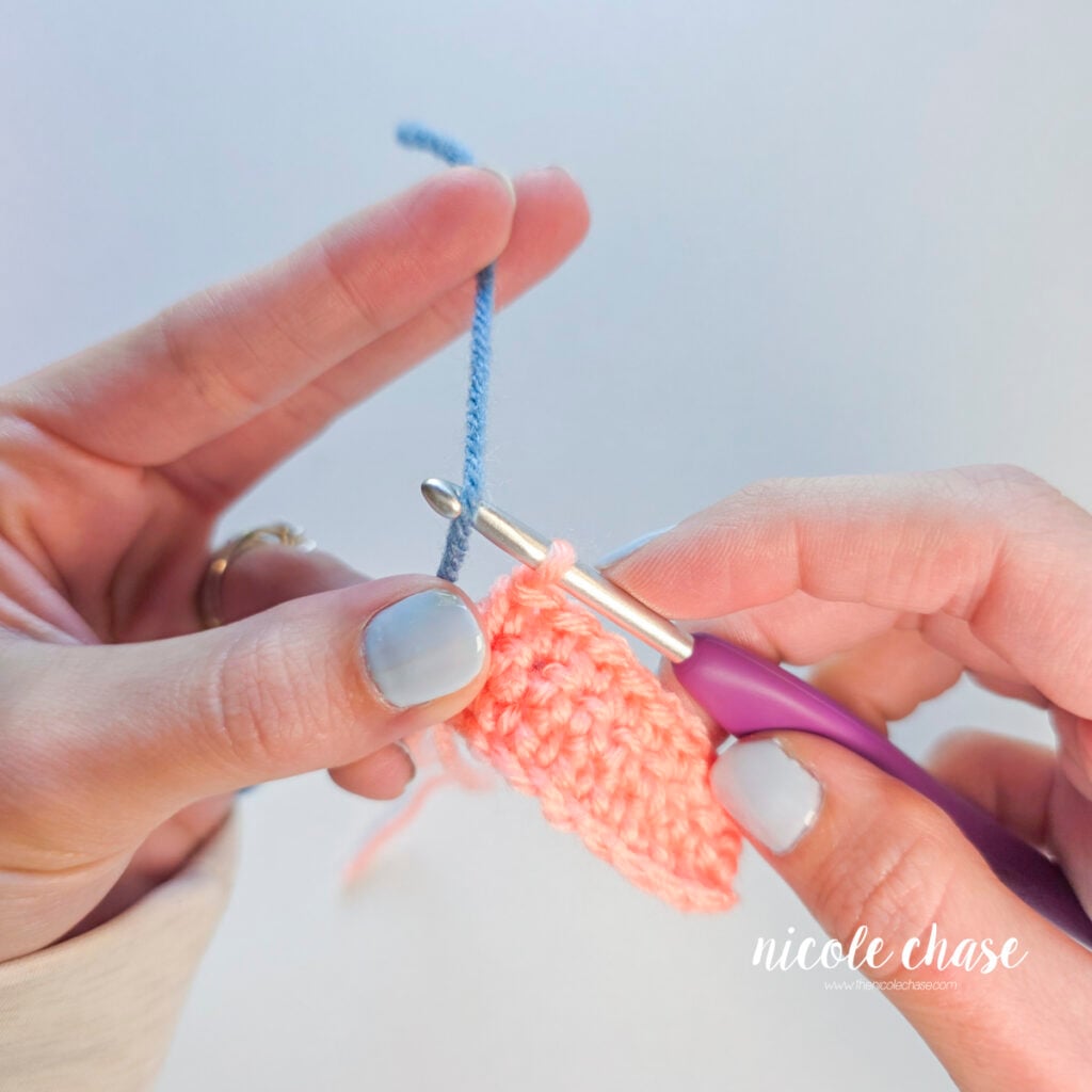 A blue yarn place onto a purple crochet hook next to a pink crochet swatch.