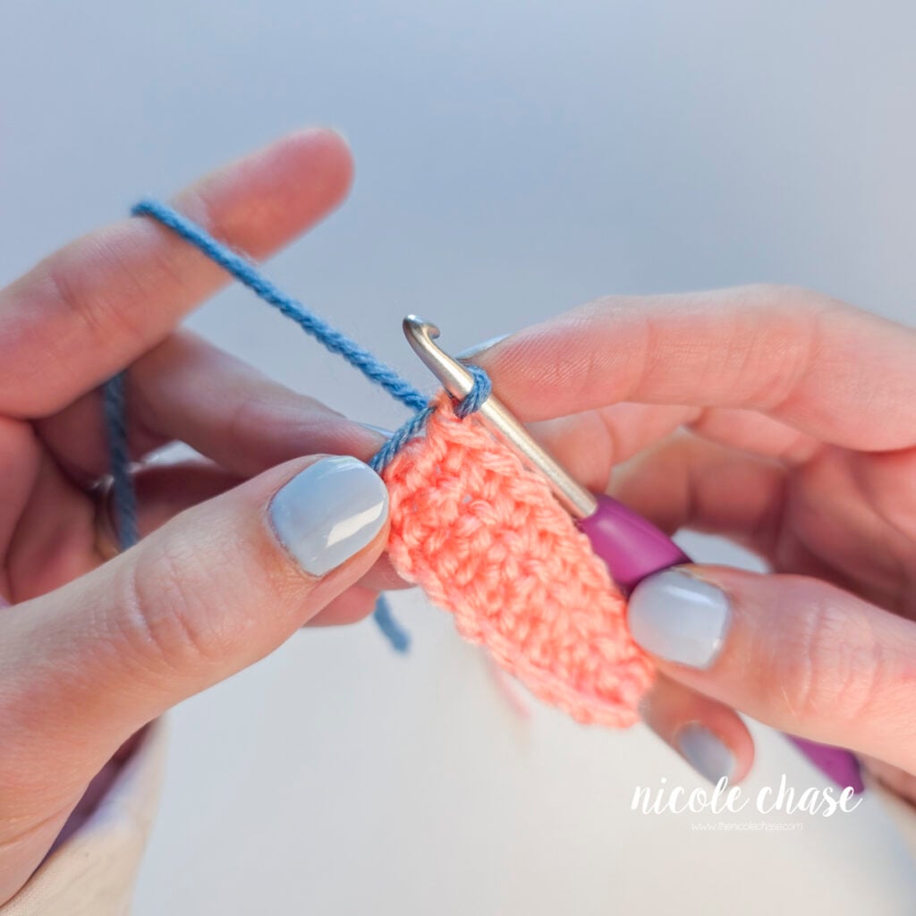 A single blue loop resting on a crochet hook at the edge of a pink crochet swatch, ready to start the next row.