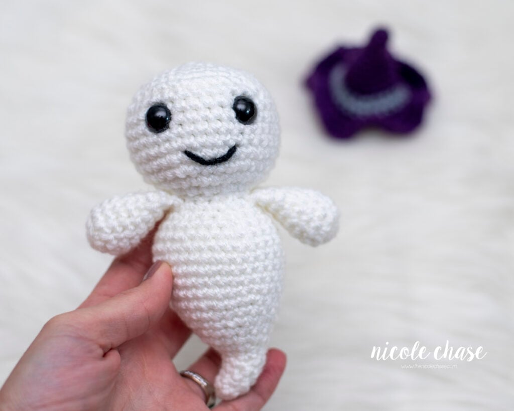 A hand holding a white crochet ghost, with a removable purple witch hat resting out of focus in the background.