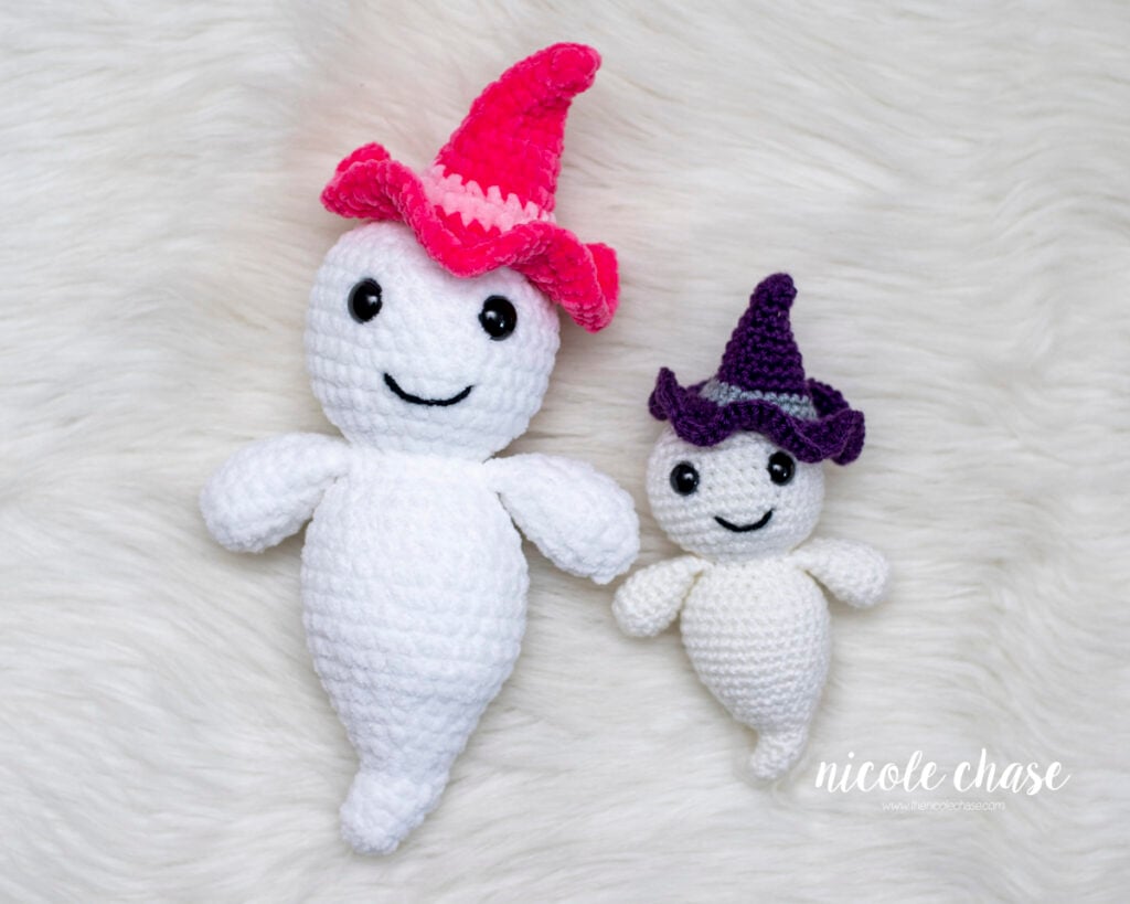 Two crocheted amigurumi ghosts named Gilbert wearing miniature witch hats side-by-side, one made with plush chenille yarn in pink and one made with worsted weight yarn in purple for the free ghost crochet pattern.