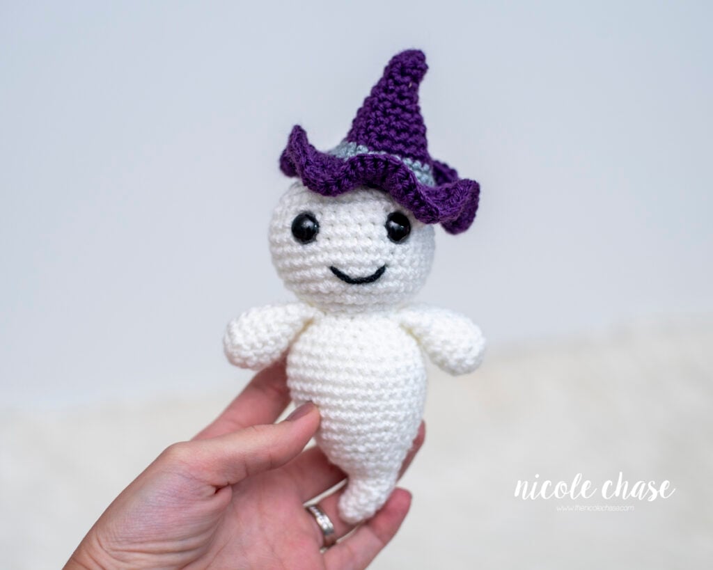 A hand holding a small crocheted amigurumi ghost named Gilbert made with white worsted weight yarn featuring safety eyes, an embroidered smile, and a purple witch hat.