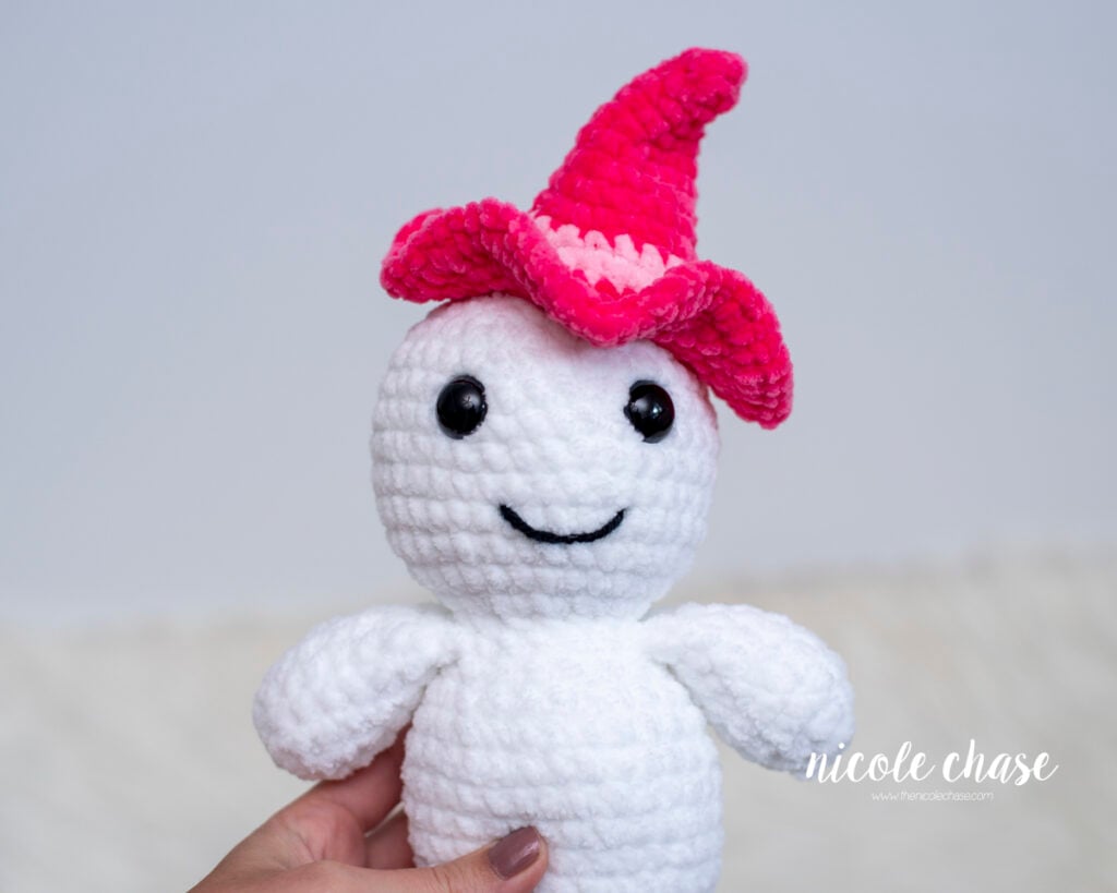 A close-up of a hand holding a white plush ghost amigurumi pattern named Gilbert made with plush chenille yarn, featuring black safety eyes, an embroidered smile, and a bright pink witch hat.