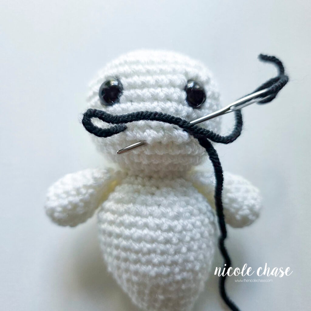 The black yarn has been looped around and the yarn needle is now coming out about one row down from the tips of the smile, where the smile will dip down.