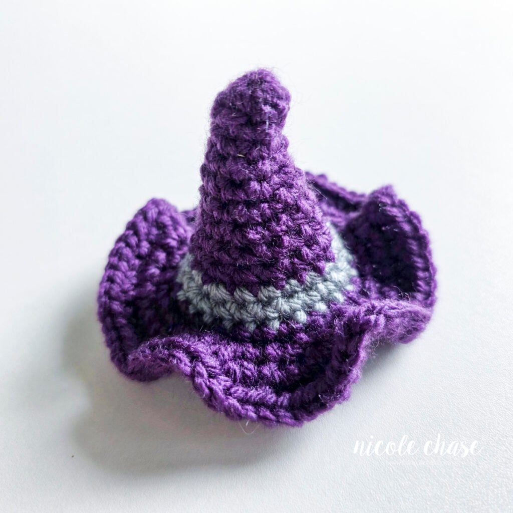 The finished witch hat made in purple yarn with a gray band. The brim is wavy and the tip of the hat is bent. The hat is ready to be placed onto the ghost amigurumi.