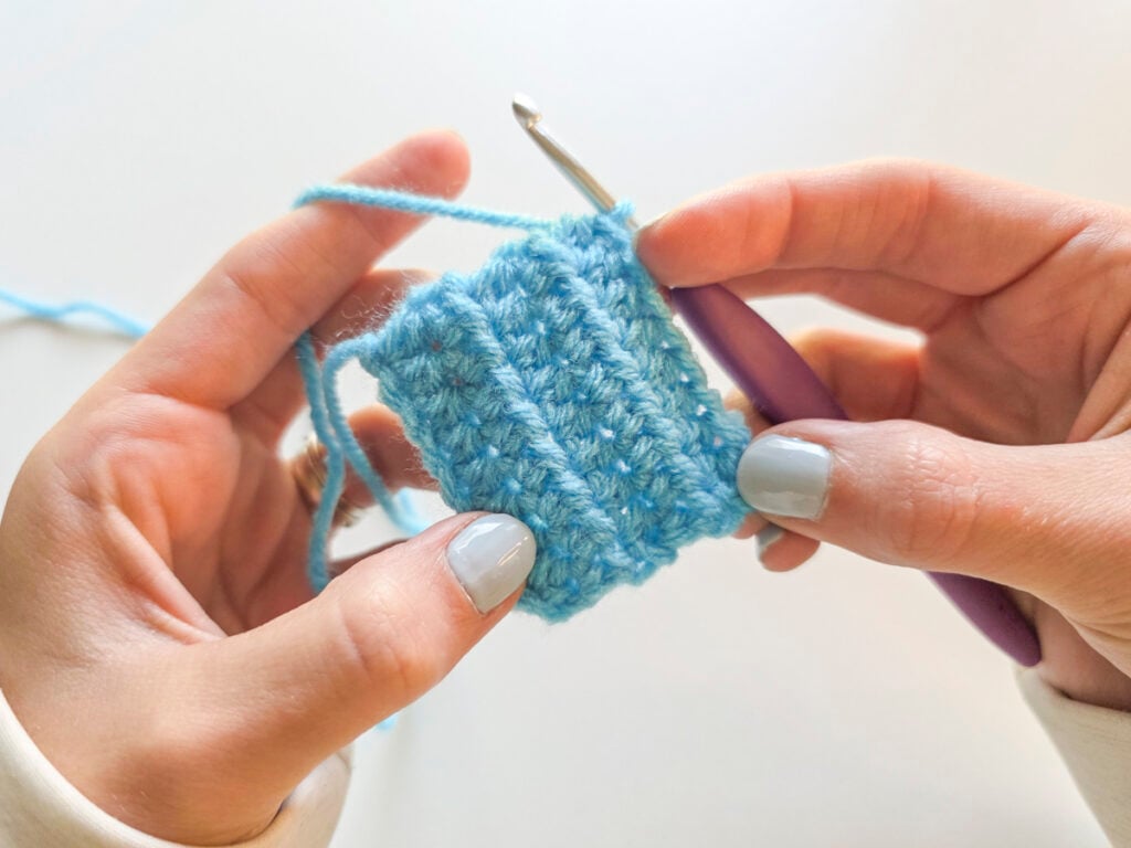Hands holding a light blue crochet swatch worked in the half double crochet stitch with a purple hook.