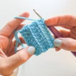 Hands holding a light blue crochet swatch worked in the half double crochet stitch with a purple hook.