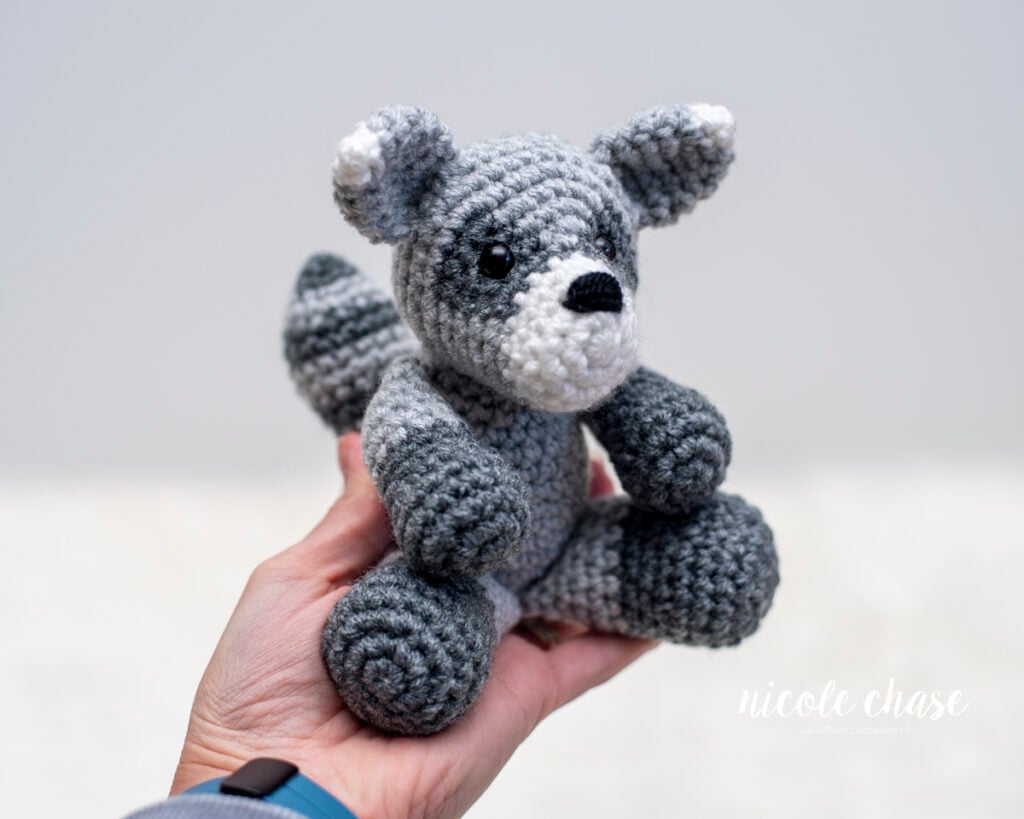 Hand holding a small crocheted raccoon amigurumi plushie to display its scale. 