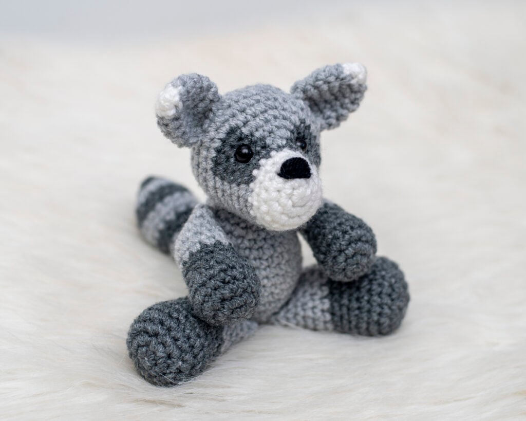 Finished amigurumi raccoon plushie made form a free raccoon crochet pattern sitting on white faux fur.