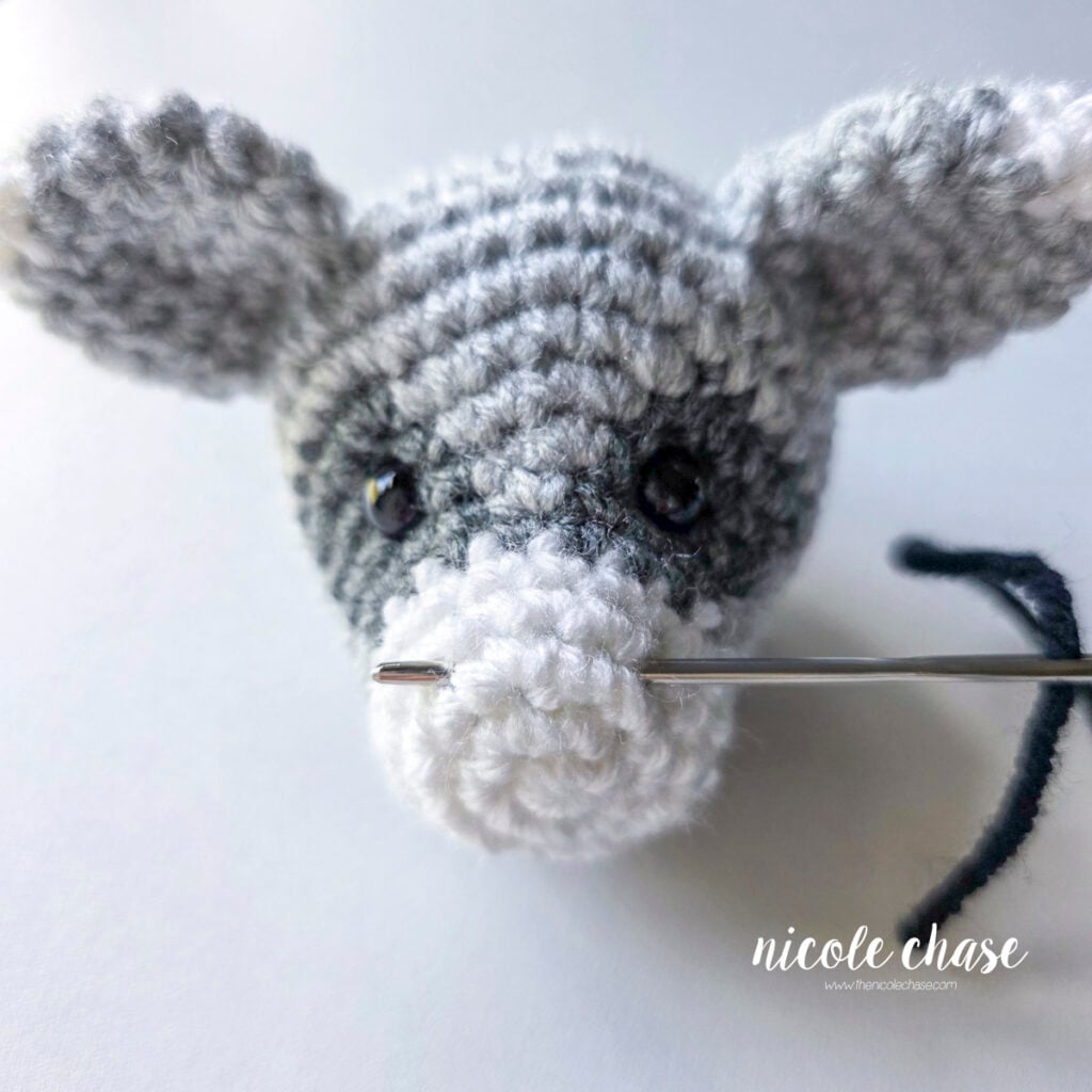 A yarn needle is placed through R3-4 of the head to start embroidering the nose on the crochet raccoon. 