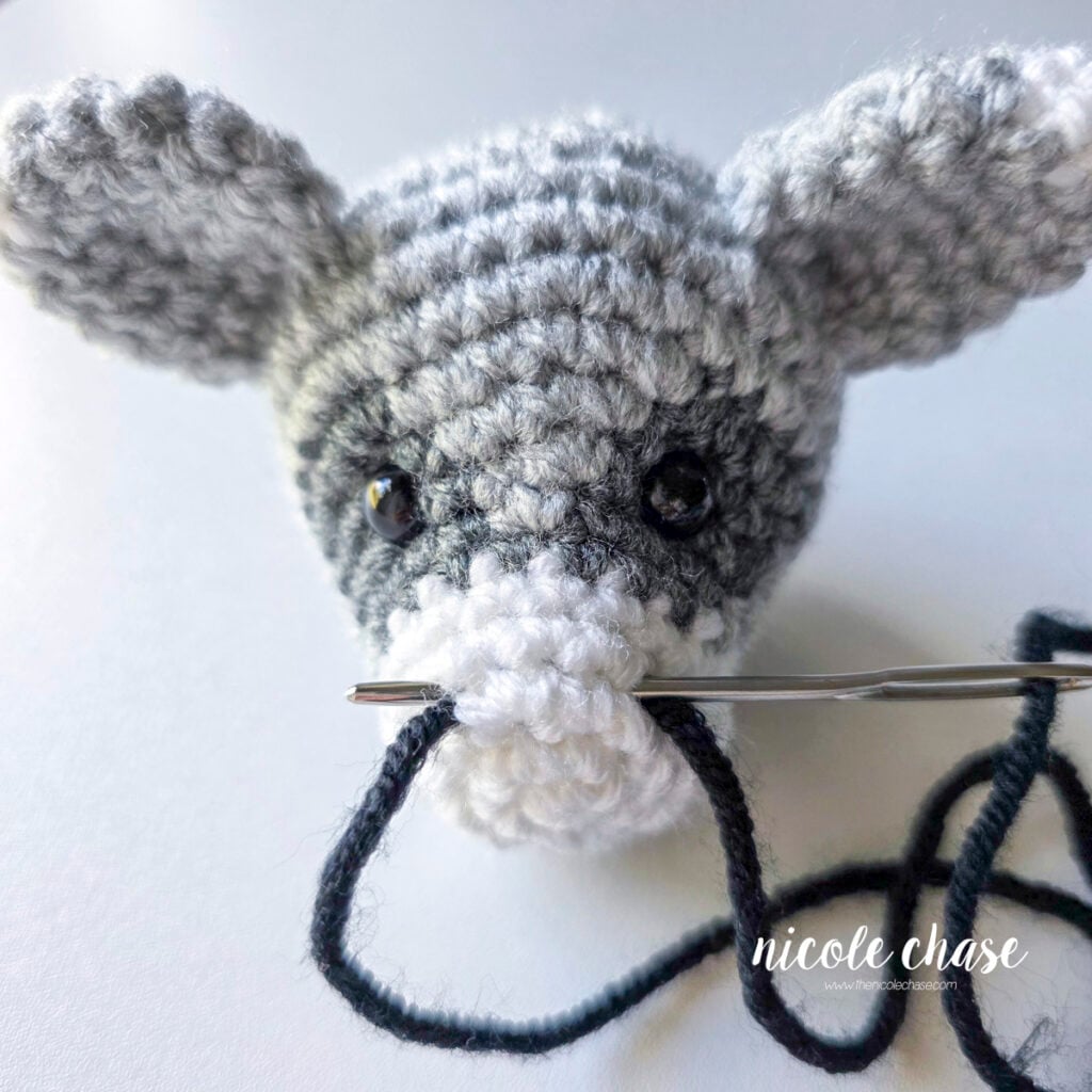 The black yarn has been looped around once and is retracing the line to embroider the nose on the crochet raccoon. 
