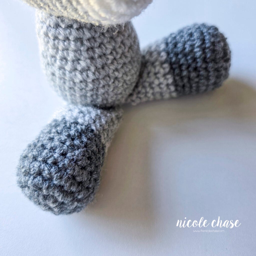 The crochet legs of the raccoon has been sewn into place on the bottom of the body, and the raccoon is in a sitting position. 