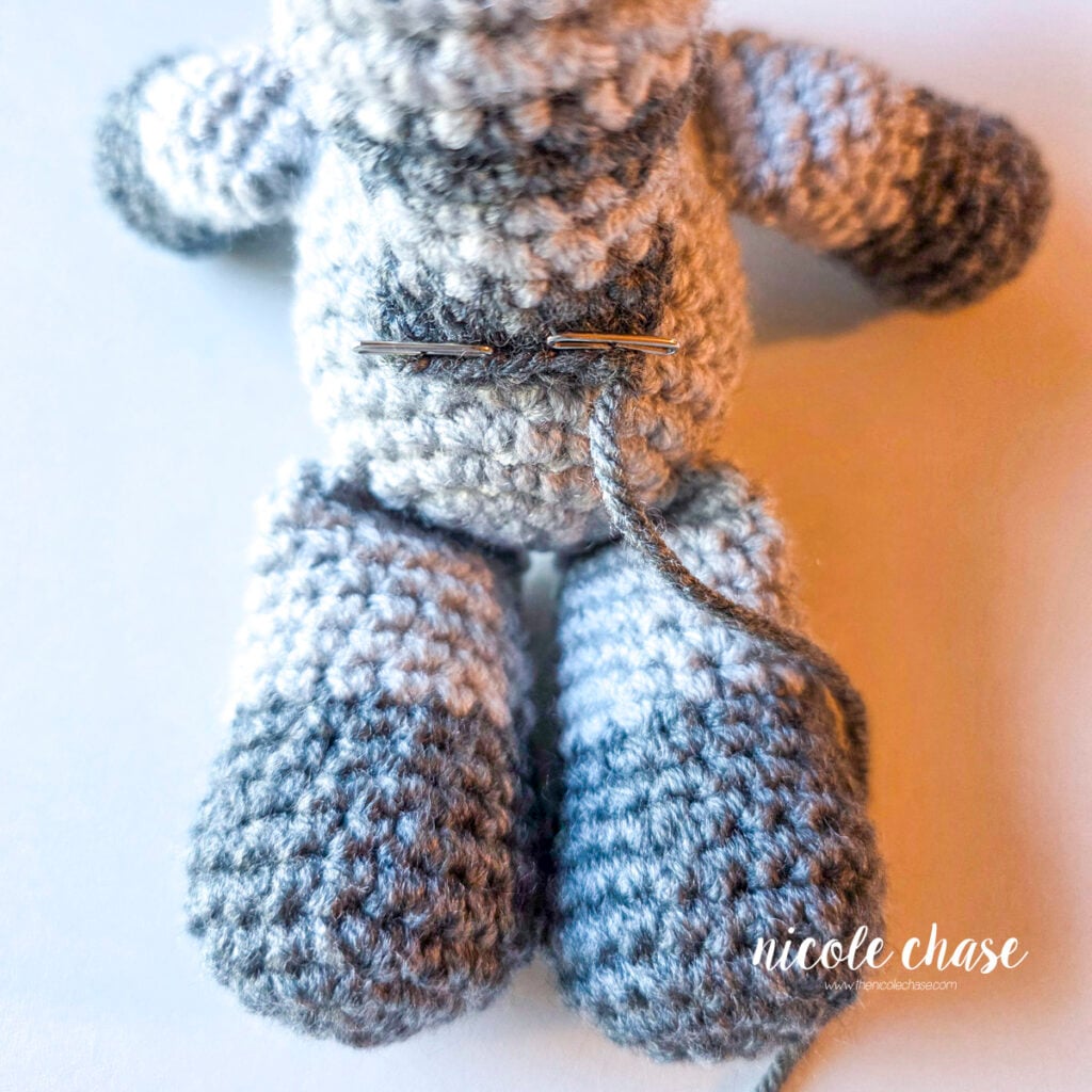 The striped tail of the crochet raccoon has been pinned into place on the back of the body and is awaiting sewing. 